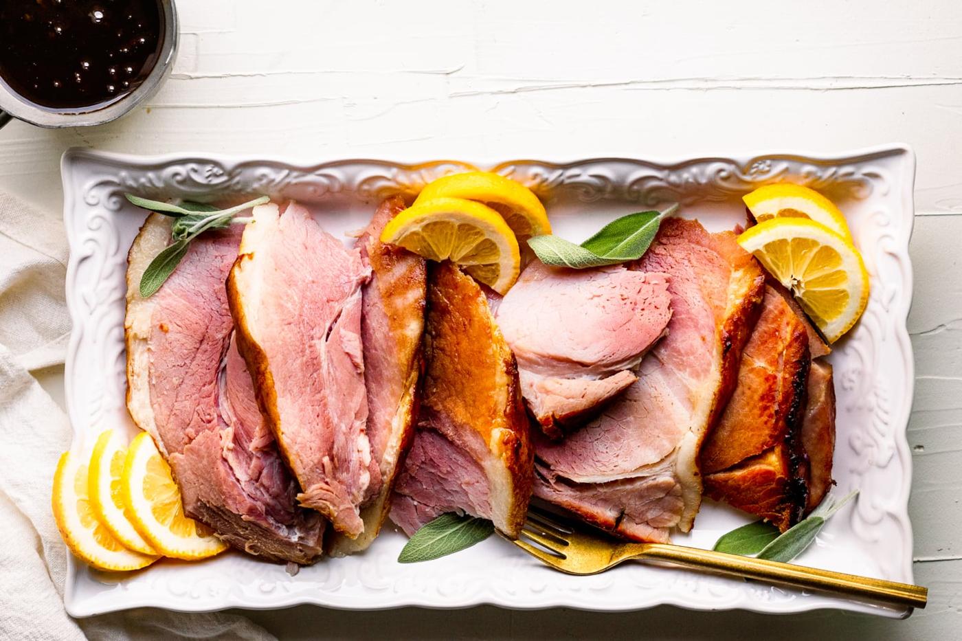 sliced baked ham on a white platter, lemon slices, and sage leaves
