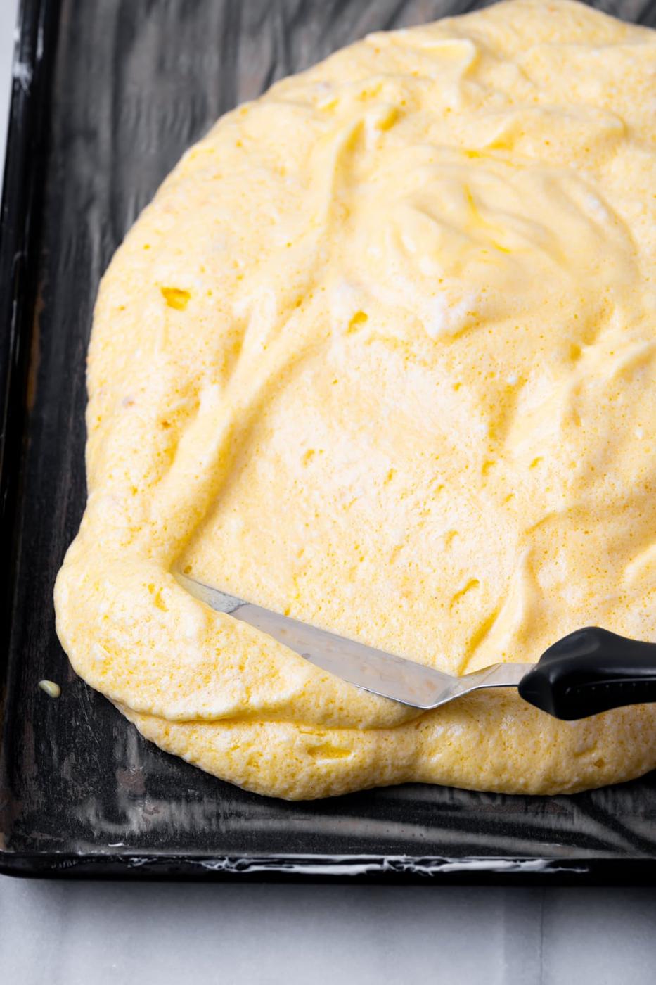 An offset spatula spreading out the batter for a brazo gitano in a buttered pan.