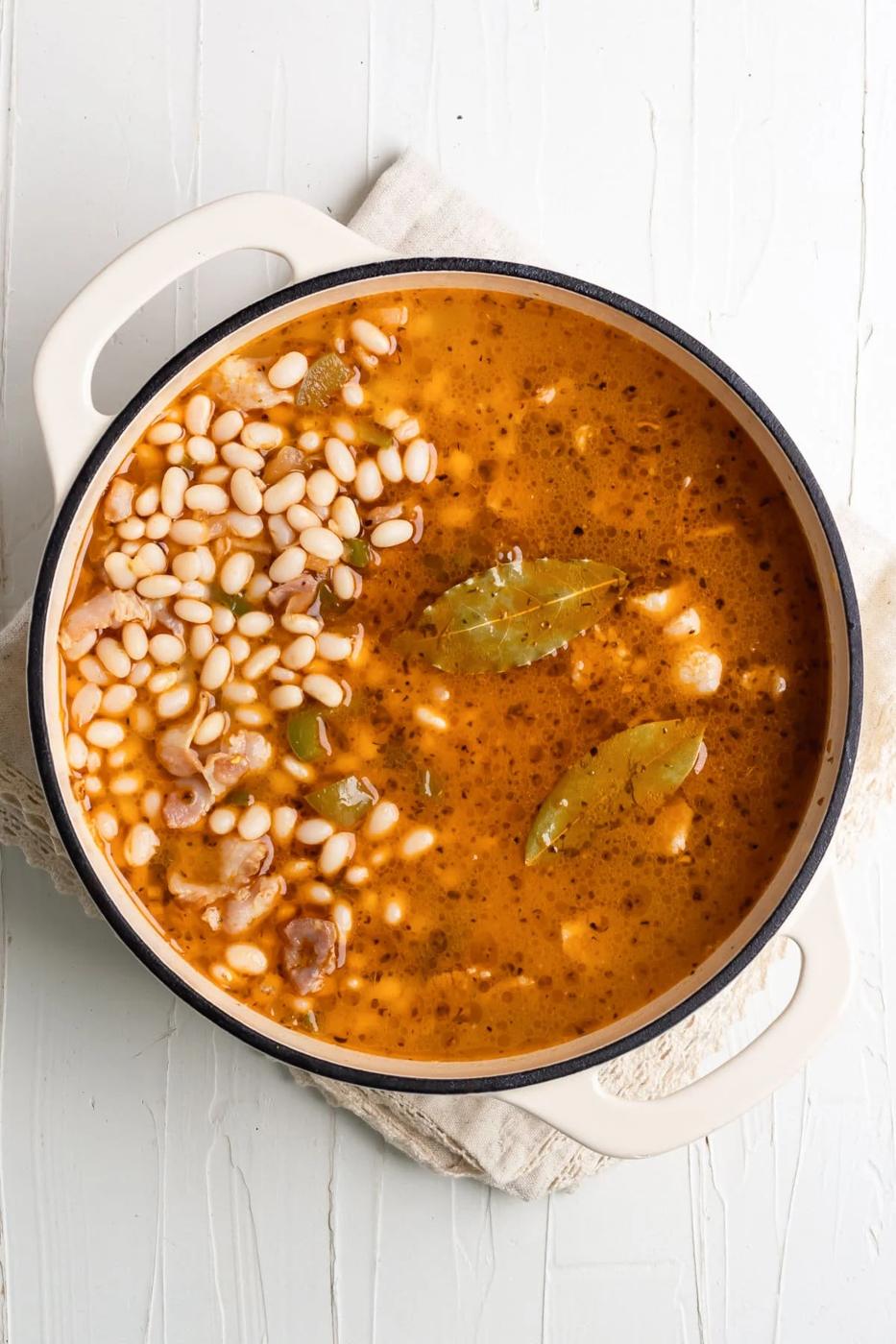 white beans in a Dutch oven cooking with spices, bay leaves, and chicken broth