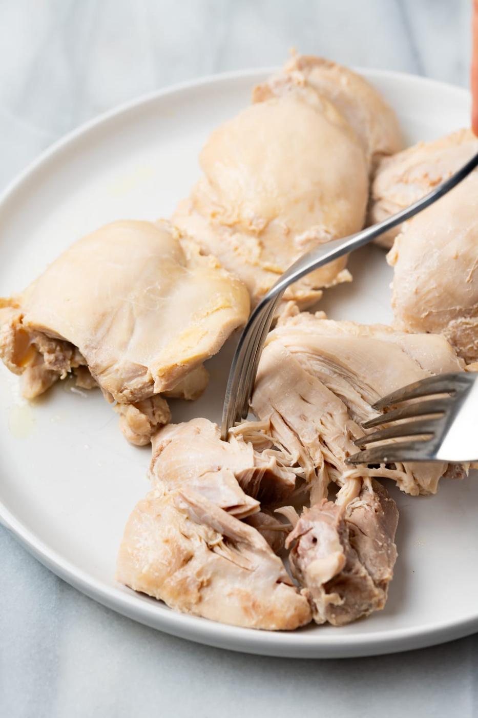 Chicken breast being shredded with two forks on a white plate with a marble background