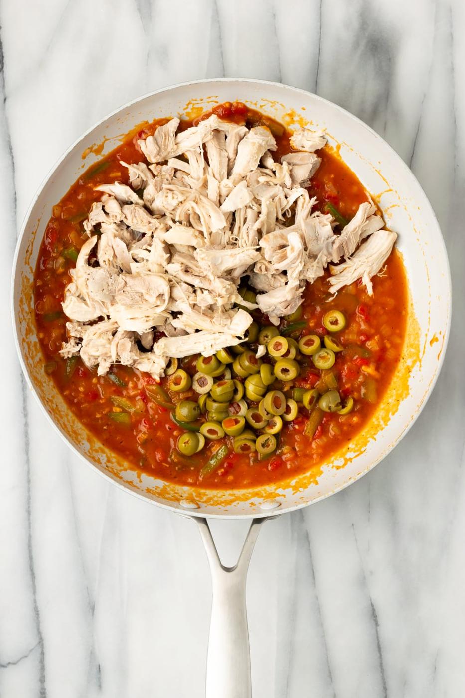 Shredded chicken and olives being added to a tomato sofrito mixture in a pan to make chicken empanada filling