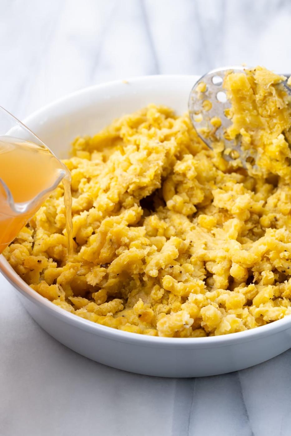 Mashed Plantains in a white bowl with a potato masher in it and a measure cup pouring in chicken broth to make fufu de platano.
