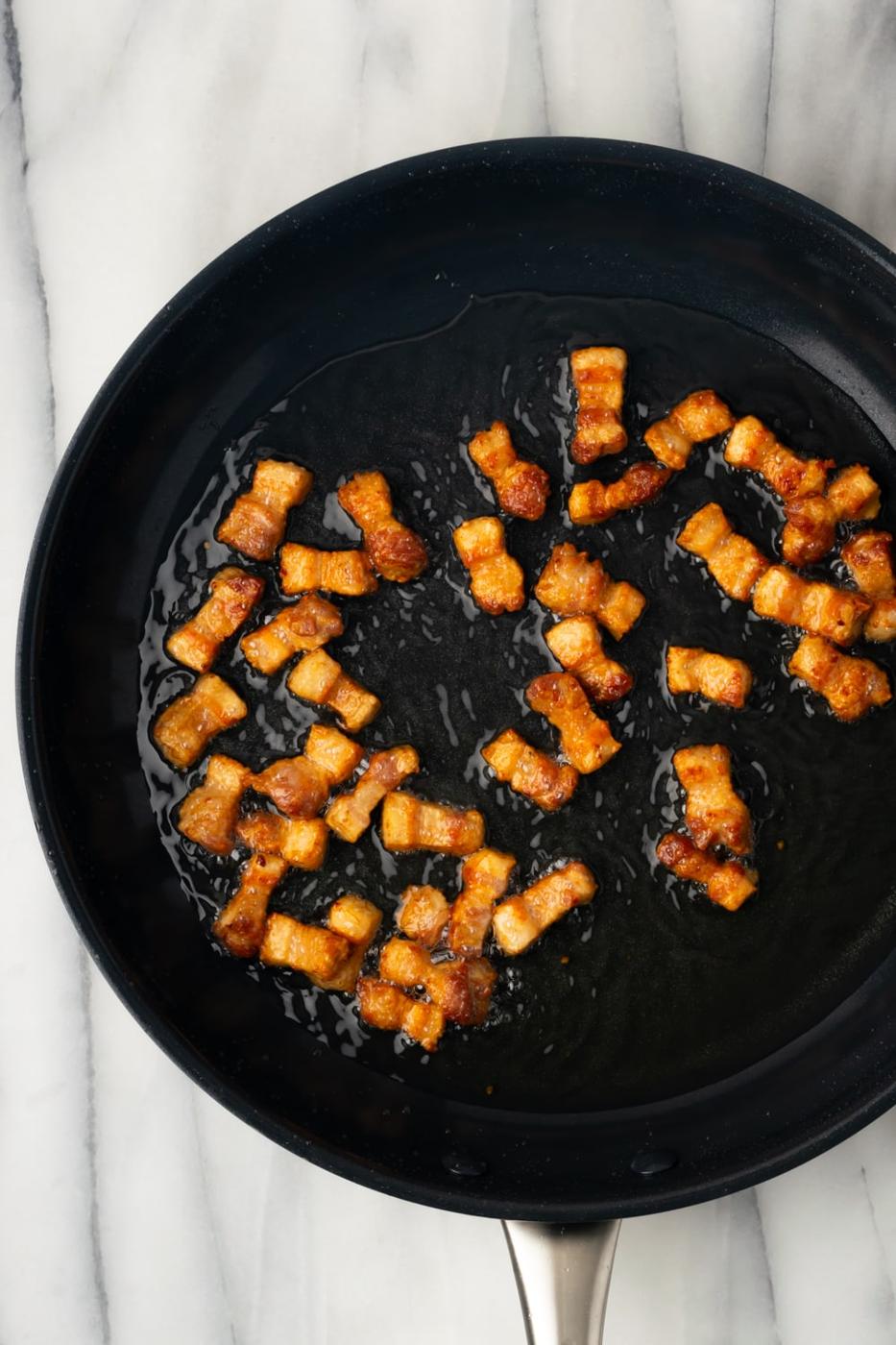 Crispy pork belly pieces in a cast iron skillet on a marble background before adding to fufu de platano.