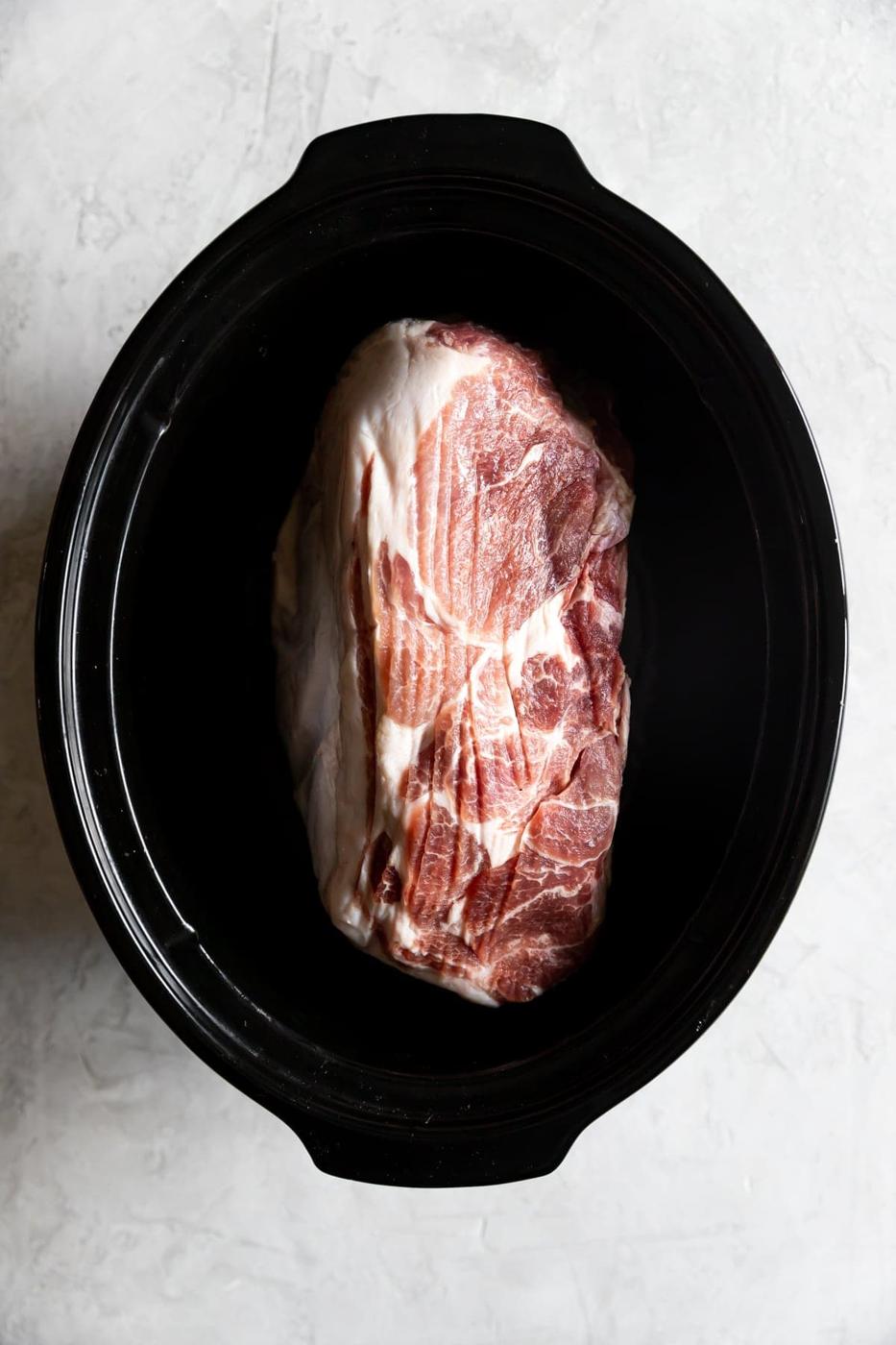A raw marbled pork shoulder, perfect for slow cooker pulled pork, sits inside a black slow cooker on a light, textured surface.