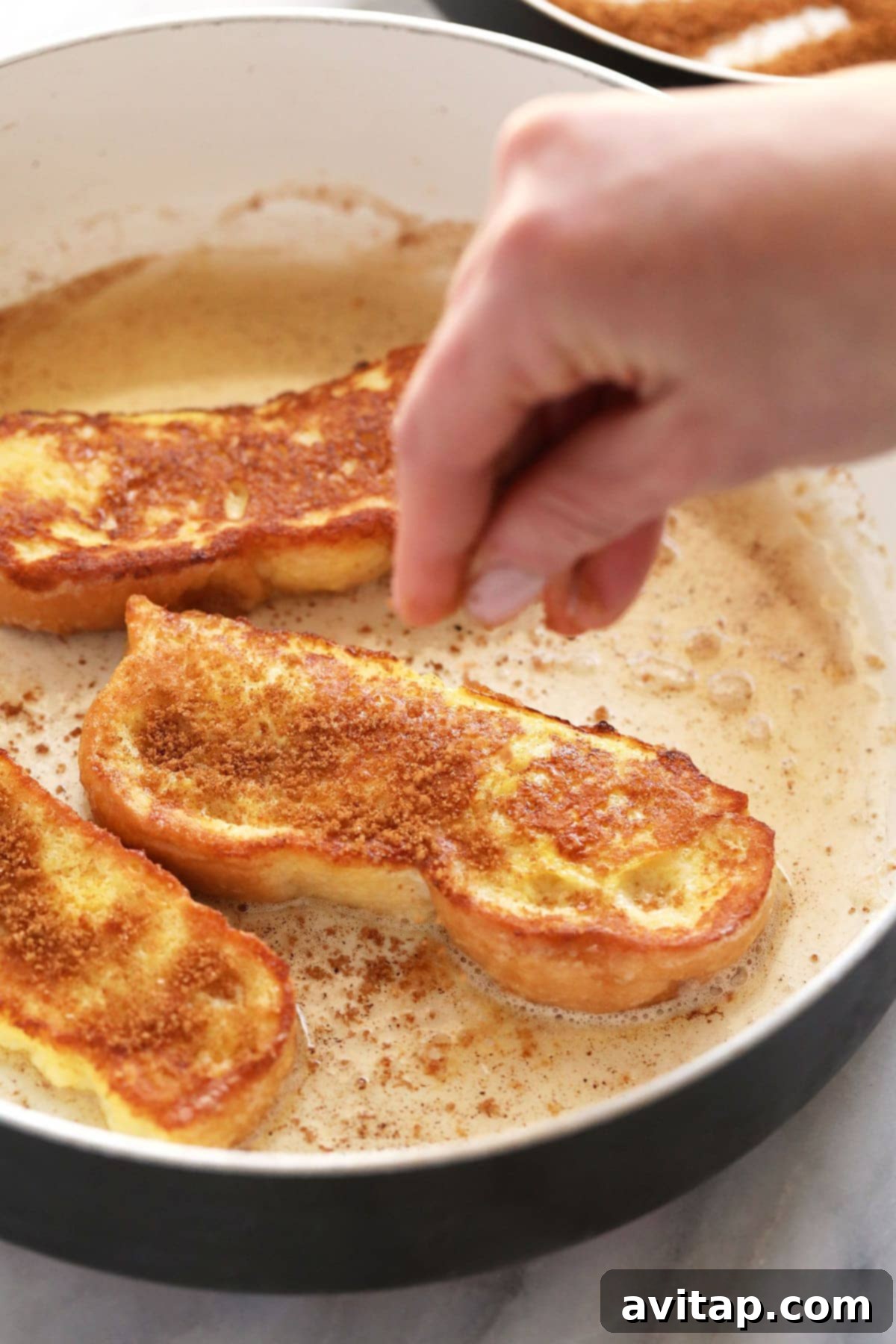 Adding Cinnamon Sugar to Hot French Toast Sticks A hand generously sprinkling a delightful amount of cinnamon sugar onto golden French Toast Sticks sizzling in a pan.