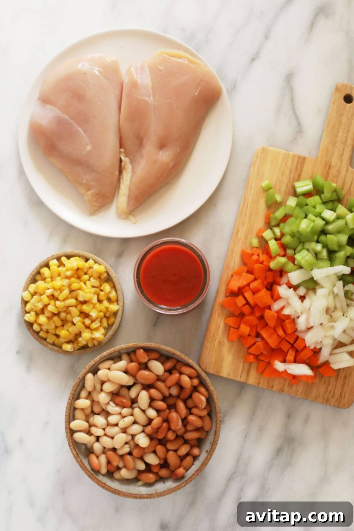 A colorful array of fresh ingredients for buffalo chicken chili laid out on a wooden cutting board, including diced vegetables, canned goods, and spices.