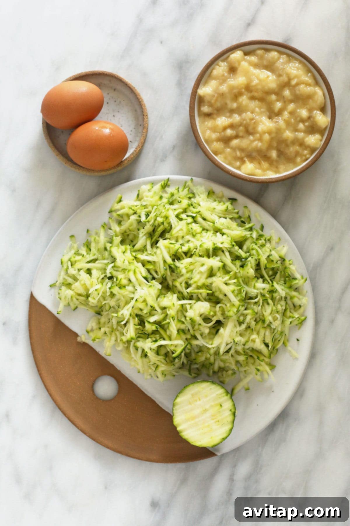 Effortless One-Bowl Zucchini Muffins 2 Plates containing shredded zucchini, cracked eggs, and mashed banana, ready for baking.