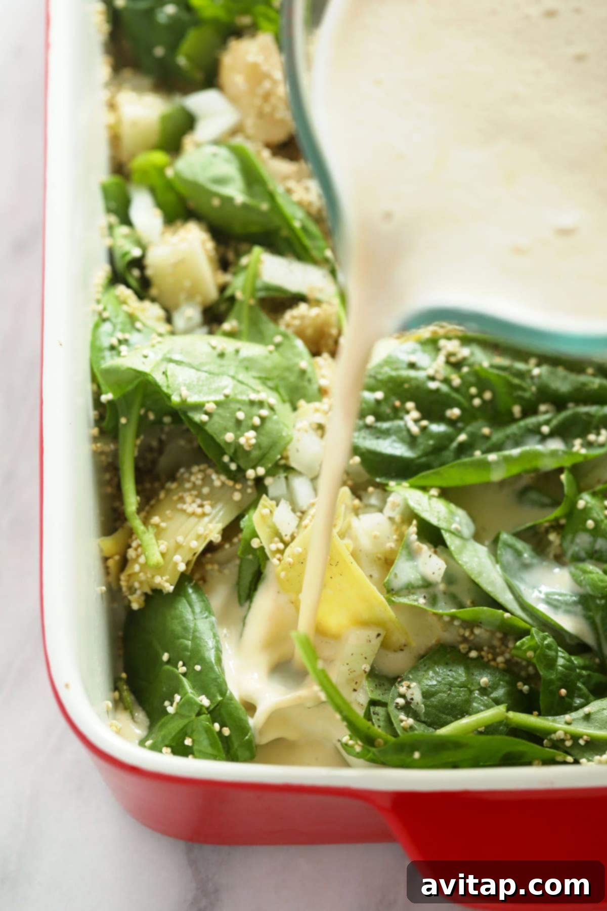 A hand pouring the creamy sauce into a casserole dish filled with quinoa, spinach, and artichokes.