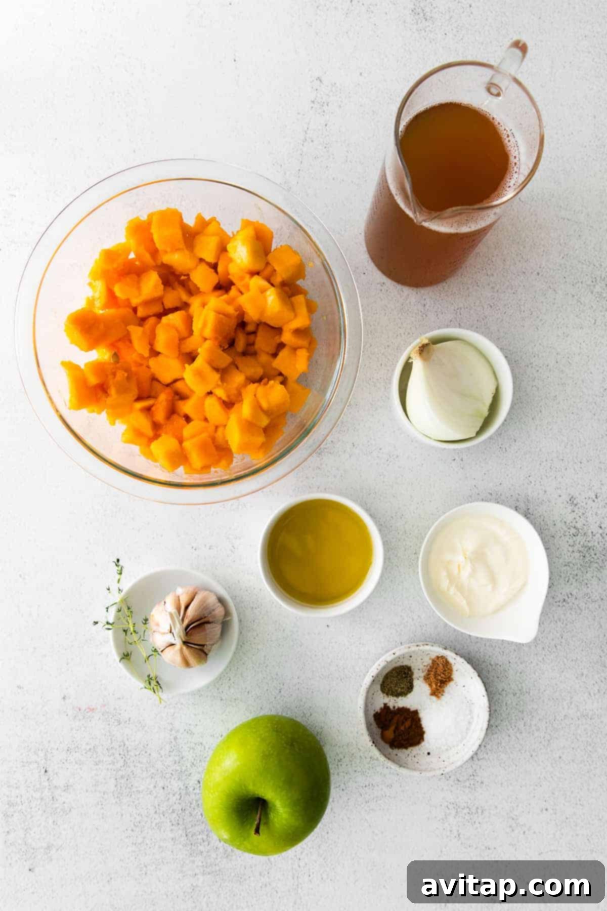 A selection of fresh, raw ingredients for butternut squash soup, including whole butternut squash, green apples, white onions, and various spices, neatly arranged on a kitchen countertop.