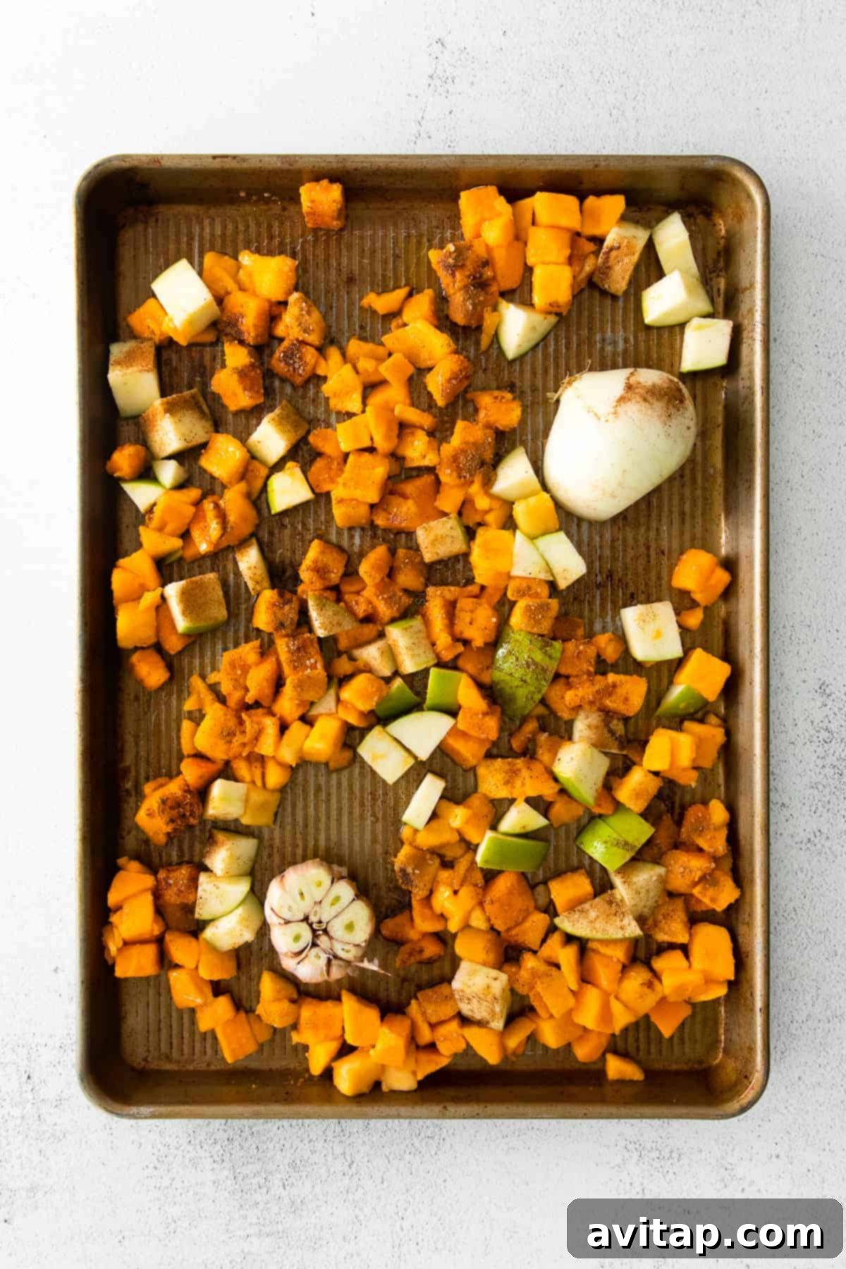 Diced butternut squash, chopped white onion, and sliced Granny Smith apple spread evenly on a baking sheet, glistening with olive oil and spices, ready for roasting.