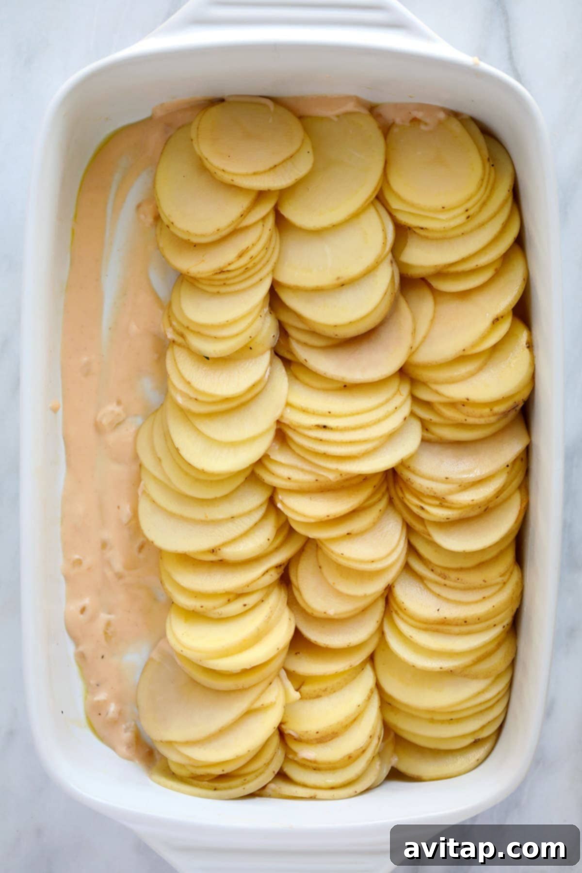 Layers of thinly sliced potatoes soaking in a creamy cheese sauce in a white ceramic baking dish, ready for the oven.