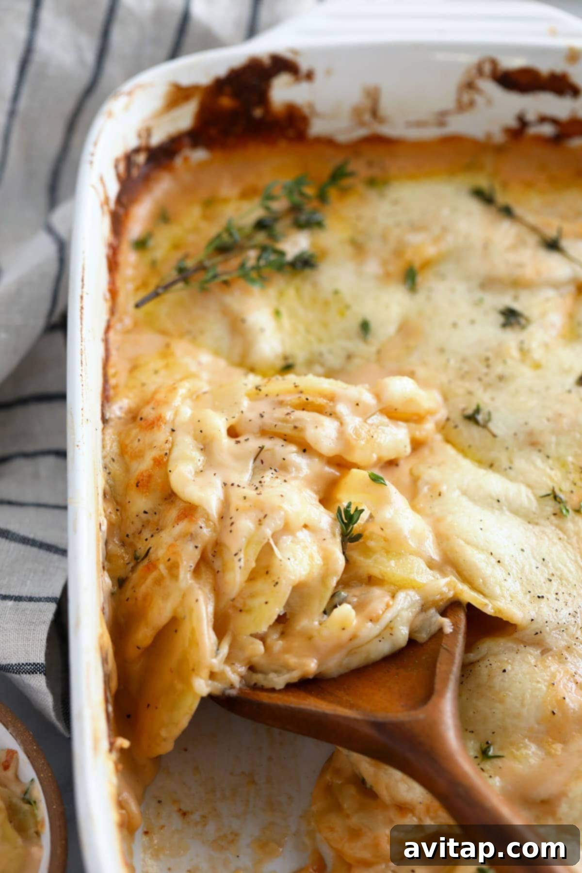 Close-up of baked cheesy scalloped potatoes in a white casserole dish, garnished with fresh thyme and showing a gooey, bubbly cheese sauce.