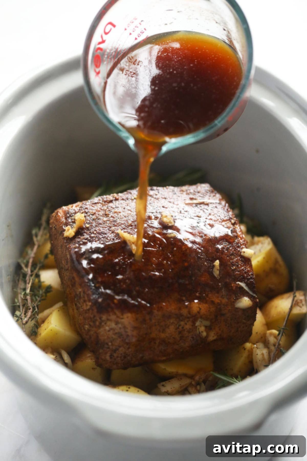A seasoned pork loin roast, nestled on a bed of fresh diced potatoes and aromatic herbs, being carefully placed into a slow cooker.