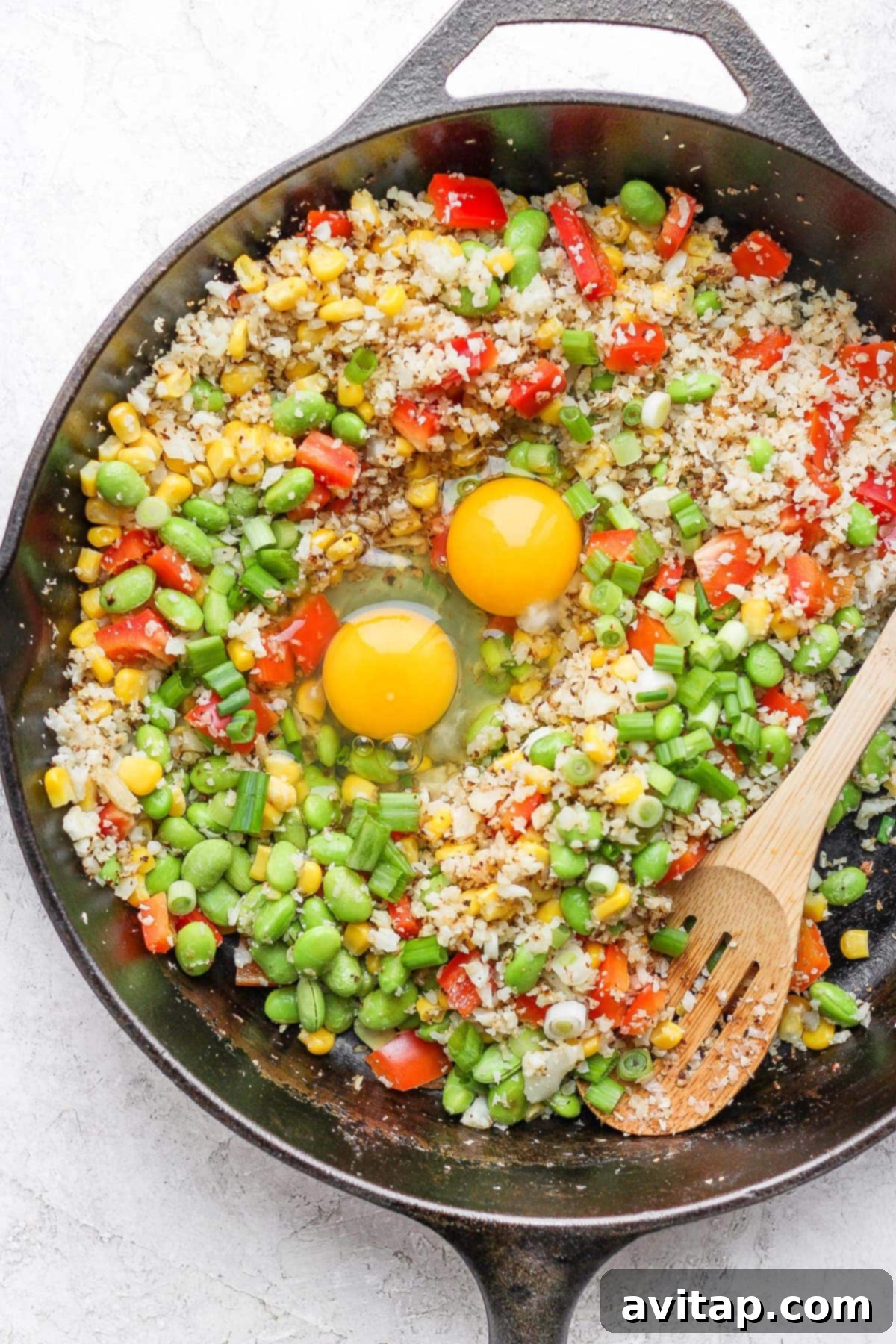 Cauliflower fried rice in a hot skillet with a well created in the center, where two eggs are cracked and ready to be scrambled.