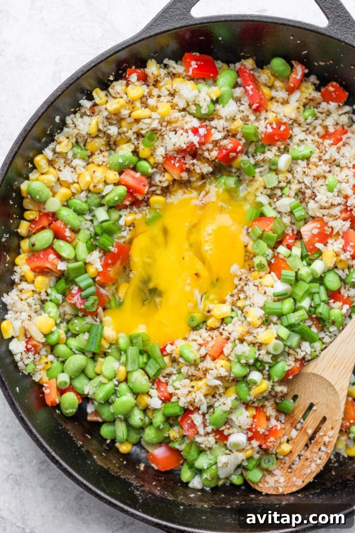 Close-up shot of cooked cauliflower fried rice with scrambled eggs, corn, edamame, and red pepper, garnished with fresh green onions in a skillet.