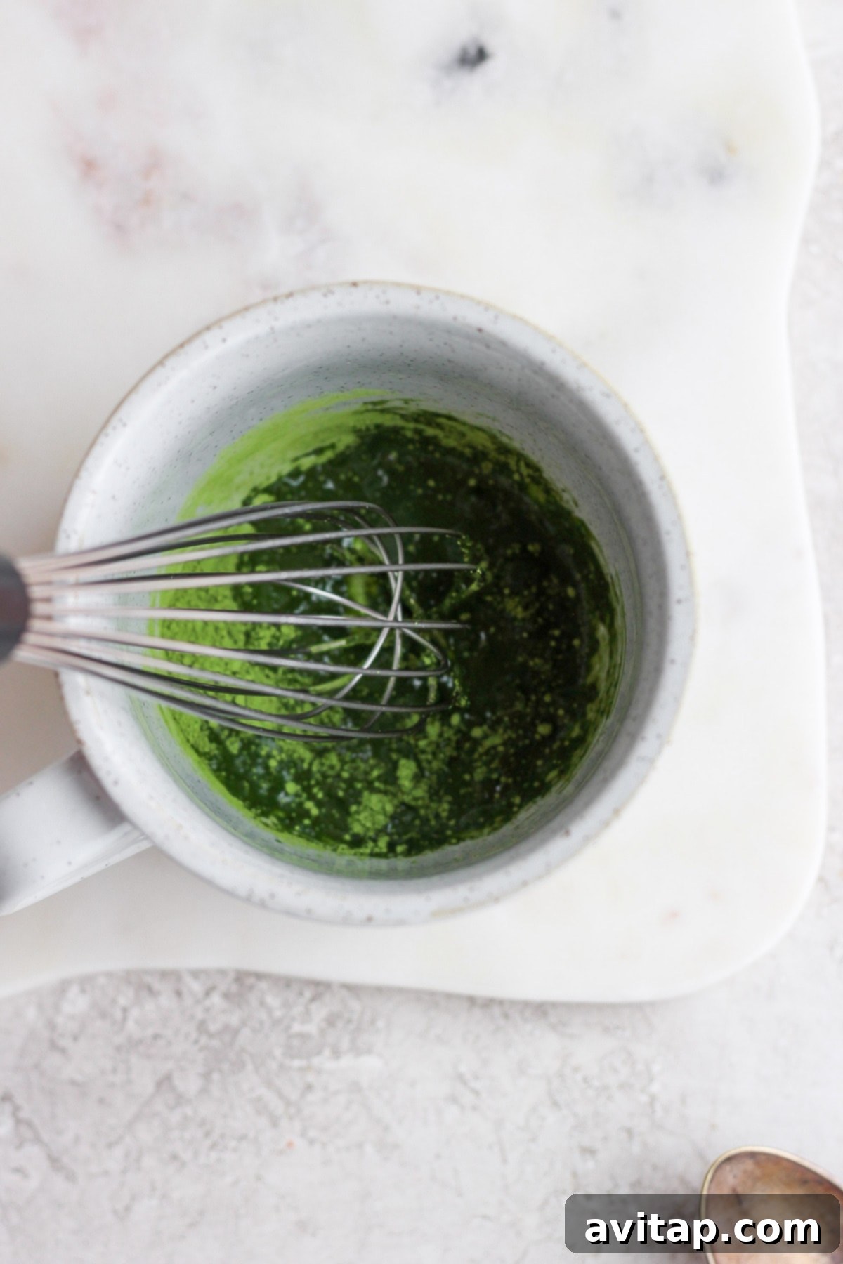 Close-up of a whisk dissolving bright green matcha powder in hot water in a mug, demonstrating the first step in making a matcha latte.