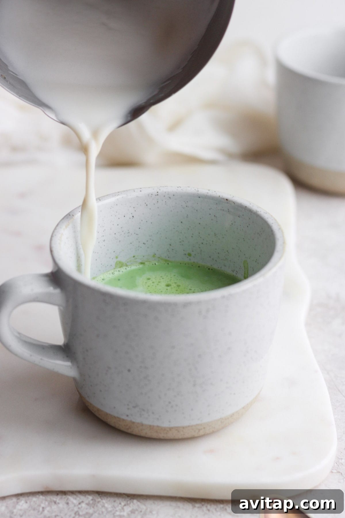A stream of frothy steamed milk being poured into a mug containing dissolved matcha powder, creating a beautiful layered effect.