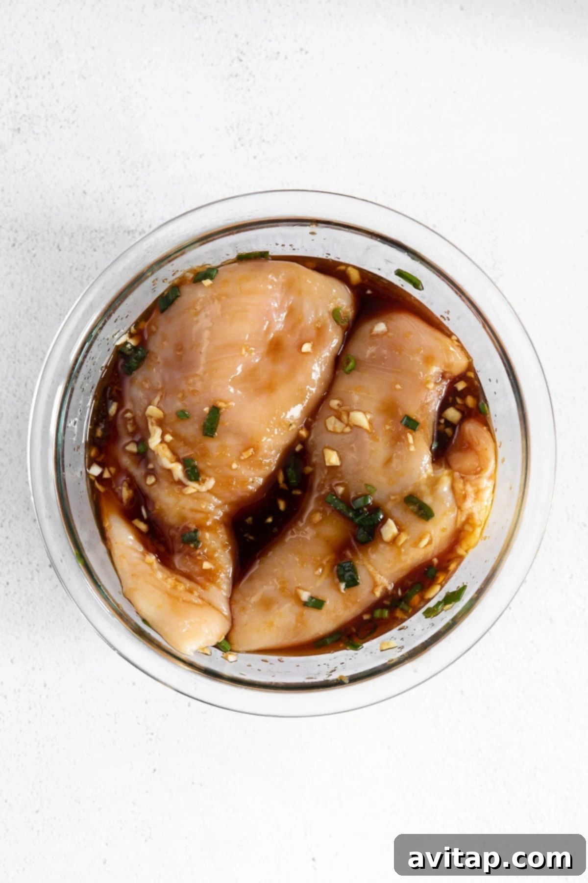 Raw chicken breast soaking in a rich, dark teriyaki marinade in a clear glass bowl, ready for baking