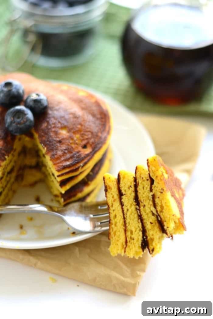 A fork piercing through a stack of golden Paleo Coconut Flour Pancakes, revealing their tender, moist interior, with a drizzle of syrup.