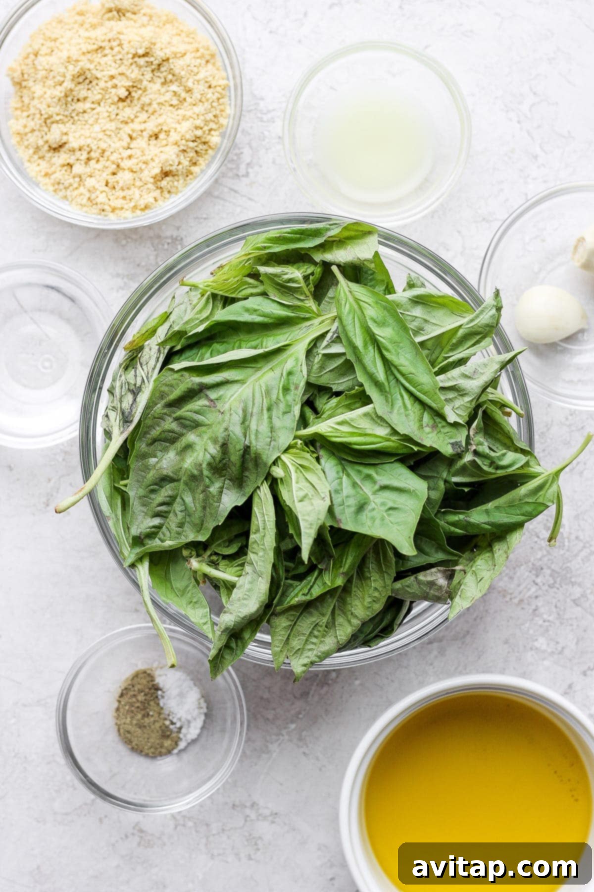 Vegan pesto is light, fresh, and 100% plant-based. It comes together with fresh basil, lemon, olive oil, garlic, and vegan parmesan cheese! Fresh basil leaves, garlic cloves, a whole lemon, homemade vegan parmesan cheese, and a bottle of olive oil arranged on a surface