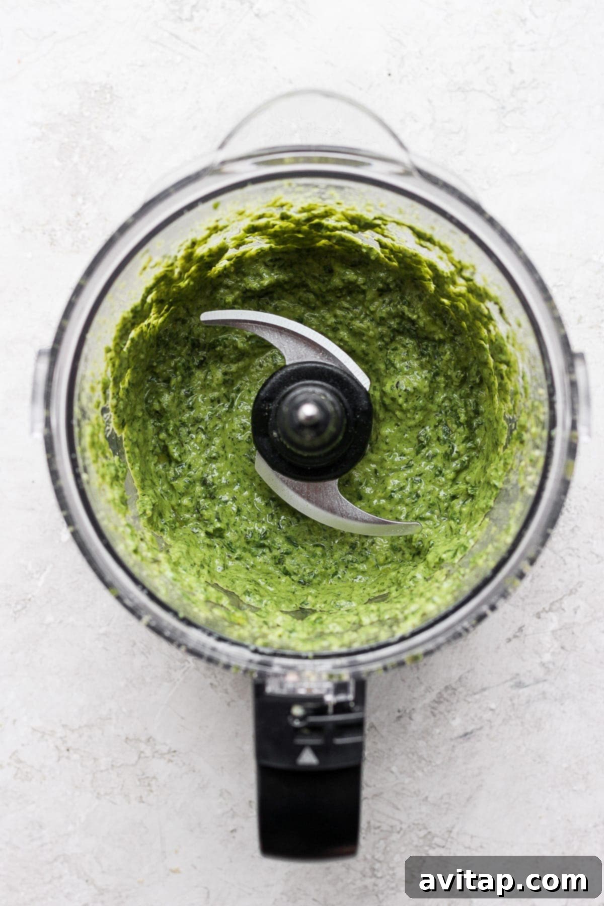 Make this simple vegan pesto recipe with fresh basil, olive oil, lemon juice, and a homemade vegan parmesan cheese. Freshly blended vegan pesto swirling inside a food processor bowl, showing its vibrant green color and smooth texture.