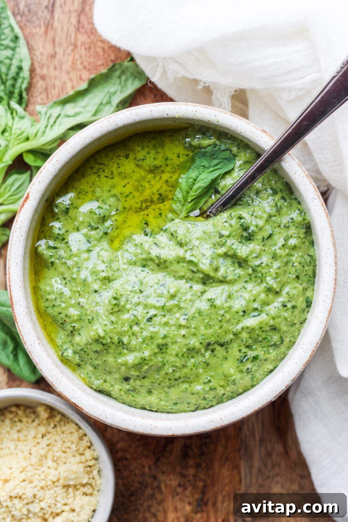 Make this simple vegan pesto recipe with fresh basil, olive oil, lemon juice, and a homemade vegan parmesan cheese. A ceramic bowl filled with vibrant green vegan pesto, garnished with fresh basil leaves, on a rustic wooden surface.