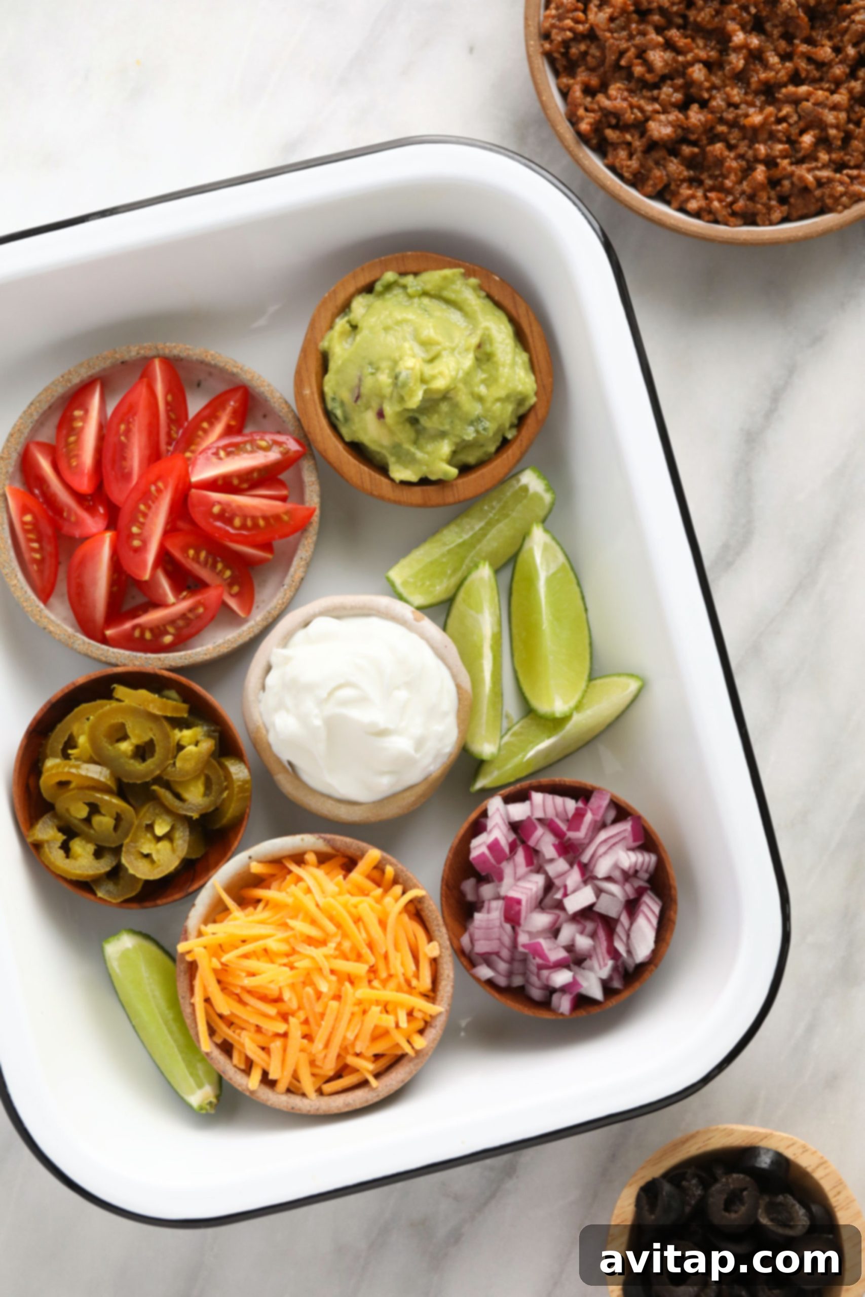 A colorful array of fresh toppings to customize your ground beef nachos. A selection of fresh, vibrant toppings for beef nachos, including sliced cherry tomatoes, black olives, jalapeños, red onion, and guacamole, in separate containers.