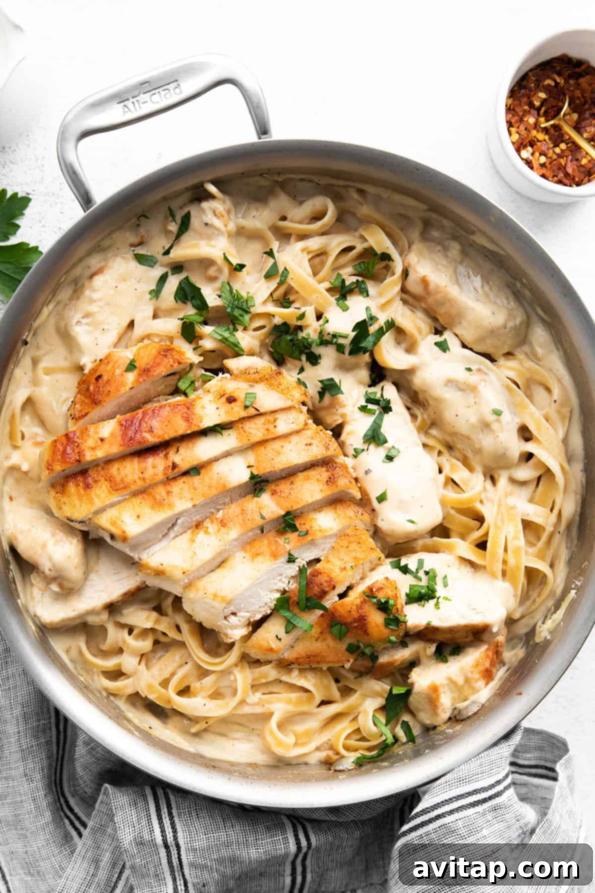 Effortless Creamy Chicken Alfredo 2 A pan filled with creamy fettuccine pasta topped with slices of golden-brown grilled chicken and garnished with fresh green parsley. In the background, a small bowl of red pepper flakes hints at an optional spice kick.