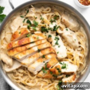Effortless Creamy Chicken Alfredo 9 A pan filled with beautifully plated creamy fettuccine pasta, generously topped with tender slices of grilled chicken and garnished with vibrant chopped parsley. A small bowl of red pepper flakes is visible in the background, offering an optional spicy touch.