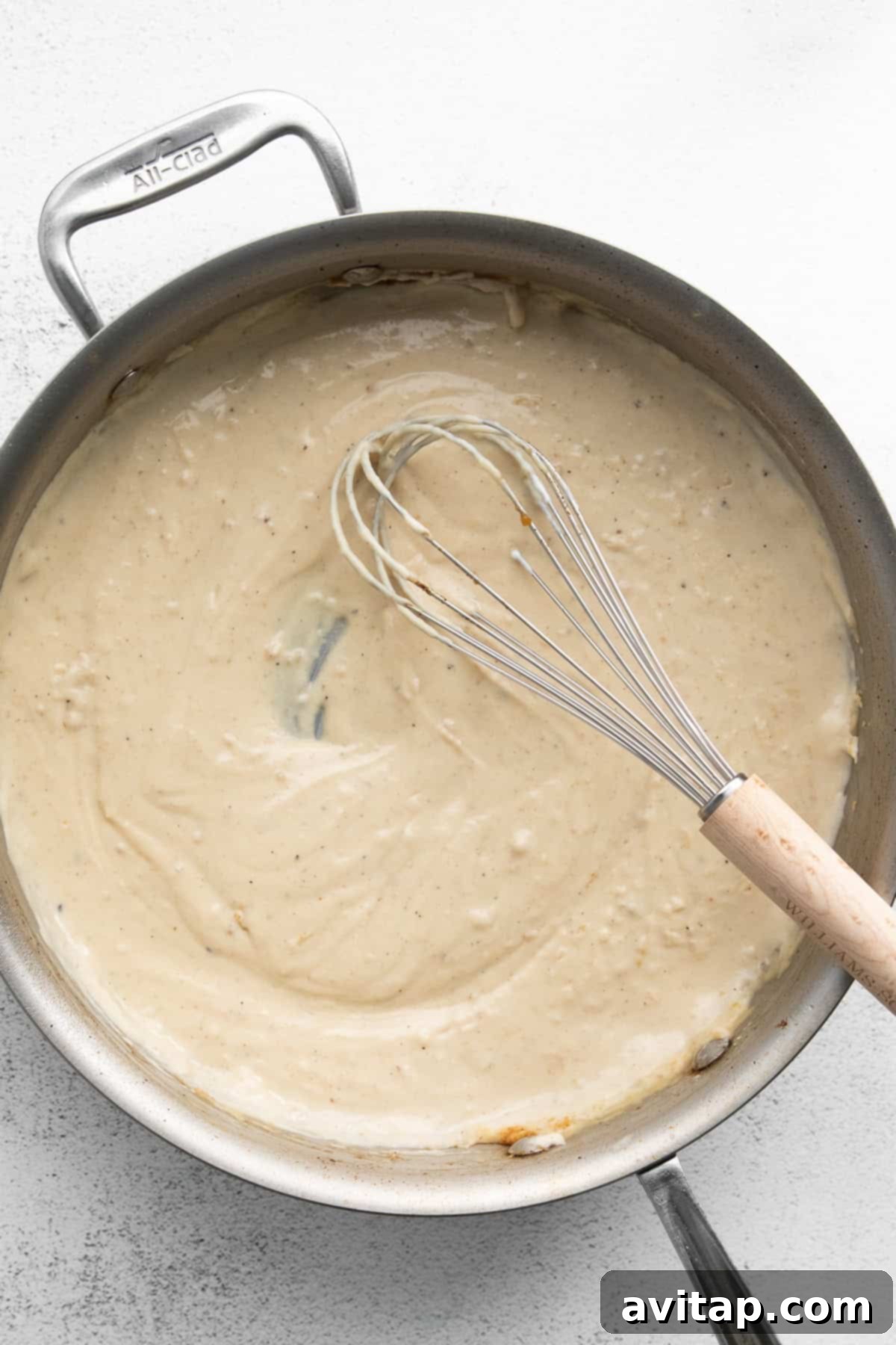 Effortless Creamy Chicken Alfredo 7 A metal whisk stirring a large, stainless steel pan filled with a creamy, rich, beige Alfredo sauce, indicating the active process of sauce preparation.