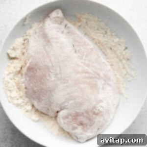 Effortless Creamy Chicken Alfredo 5 A raw chicken breast completely coated in a seasoned flour mixture, ready for pan-searing, placed on a white plate.