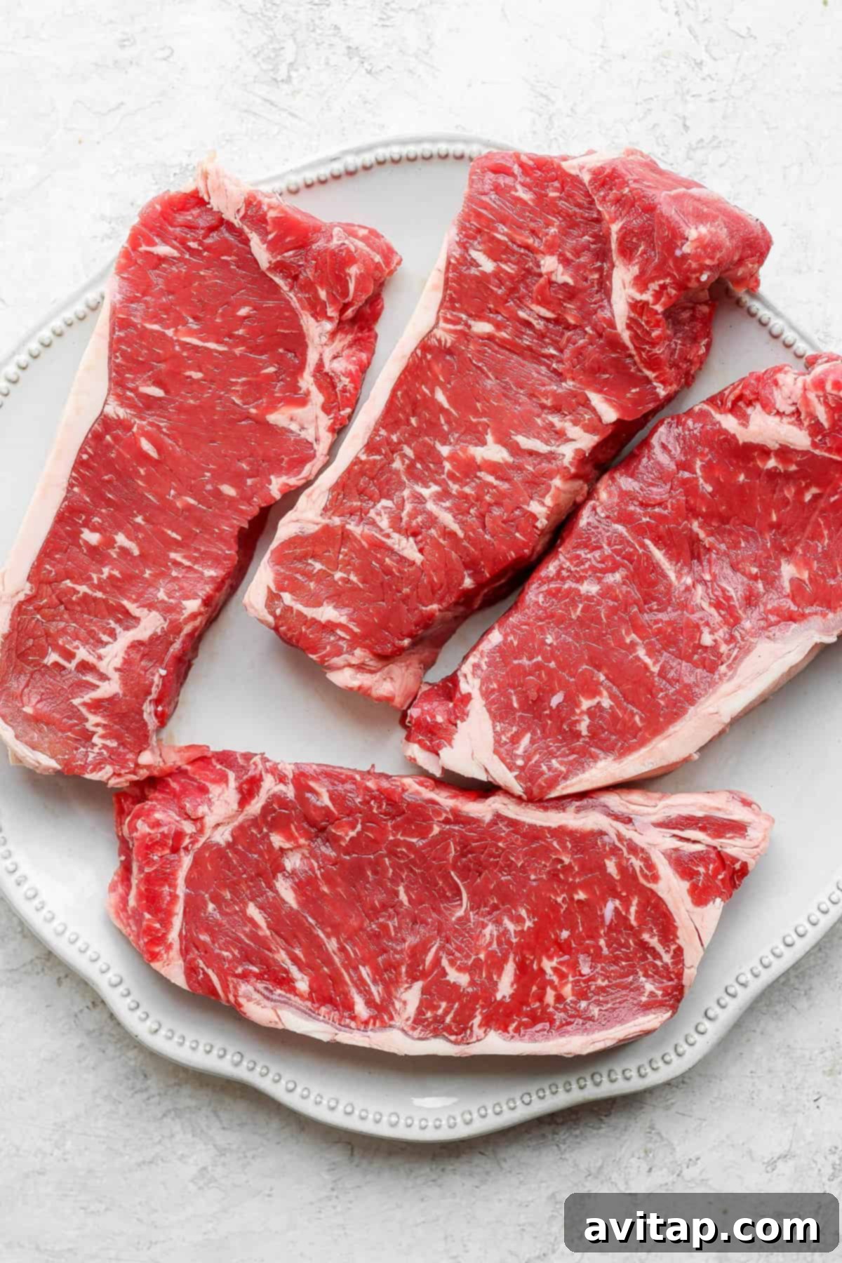 Four raw, thick-cut New York strip steaks, seasoned and ready for the grill, laid out on a plate.