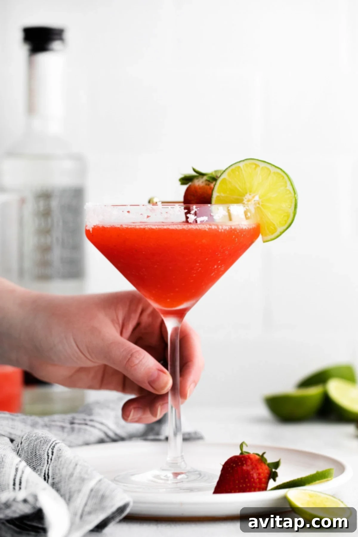 Hand holding a vibrant red frozen strawberry margarita in a salt-rimmed glass, garnished with a fresh lime wedge