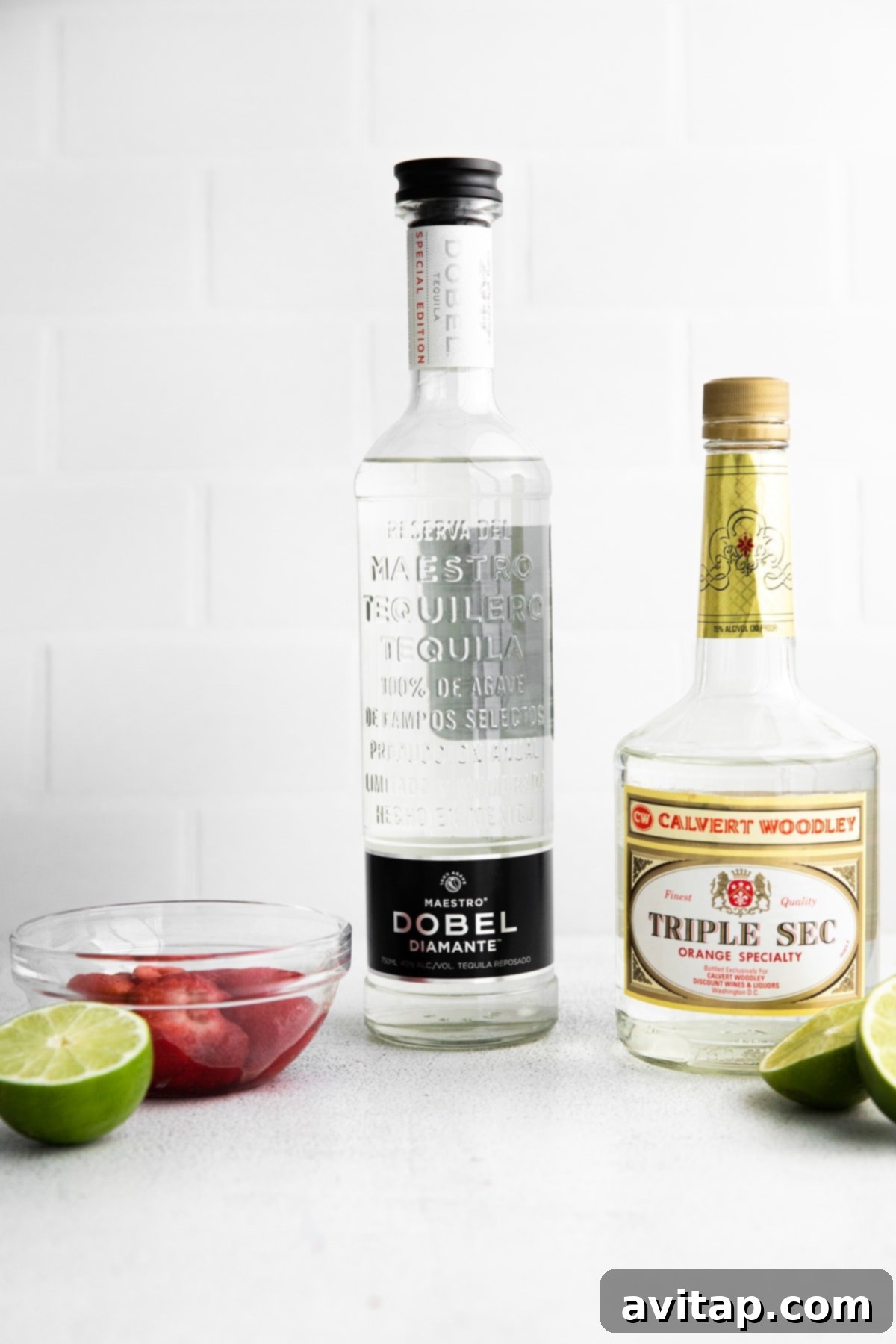 Fresh ingredients laid out for a frozen strawberry margarita, including a bottle of tequila, triple sec, a lime, and a bag of frozen strawberries