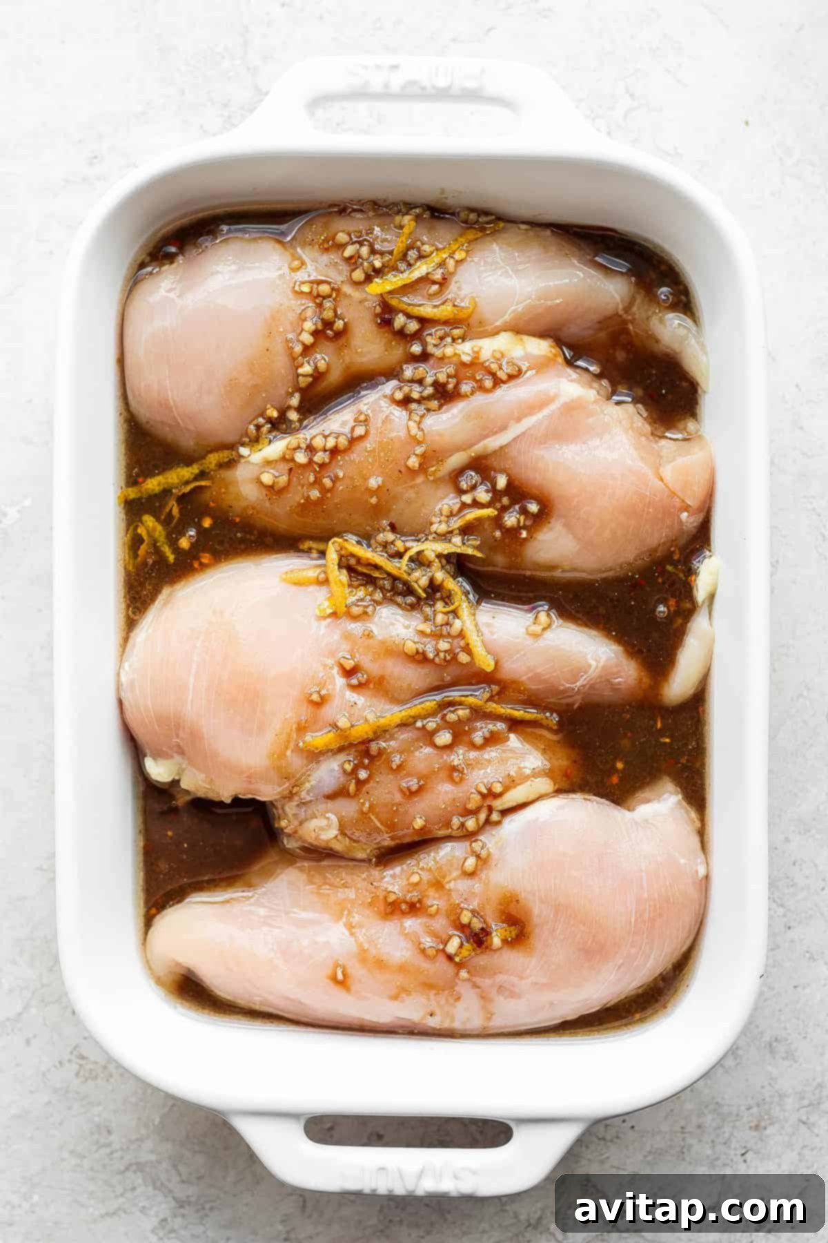 Raw chicken breasts marinating in a savory balsamic and lemon dressing inside a glass dish.