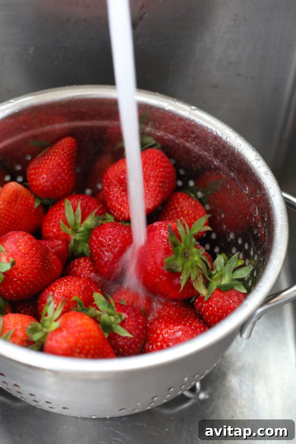 Rinsing fresh, vibrant red strawberries under cool water in a colander, preparing them for the freezing process.