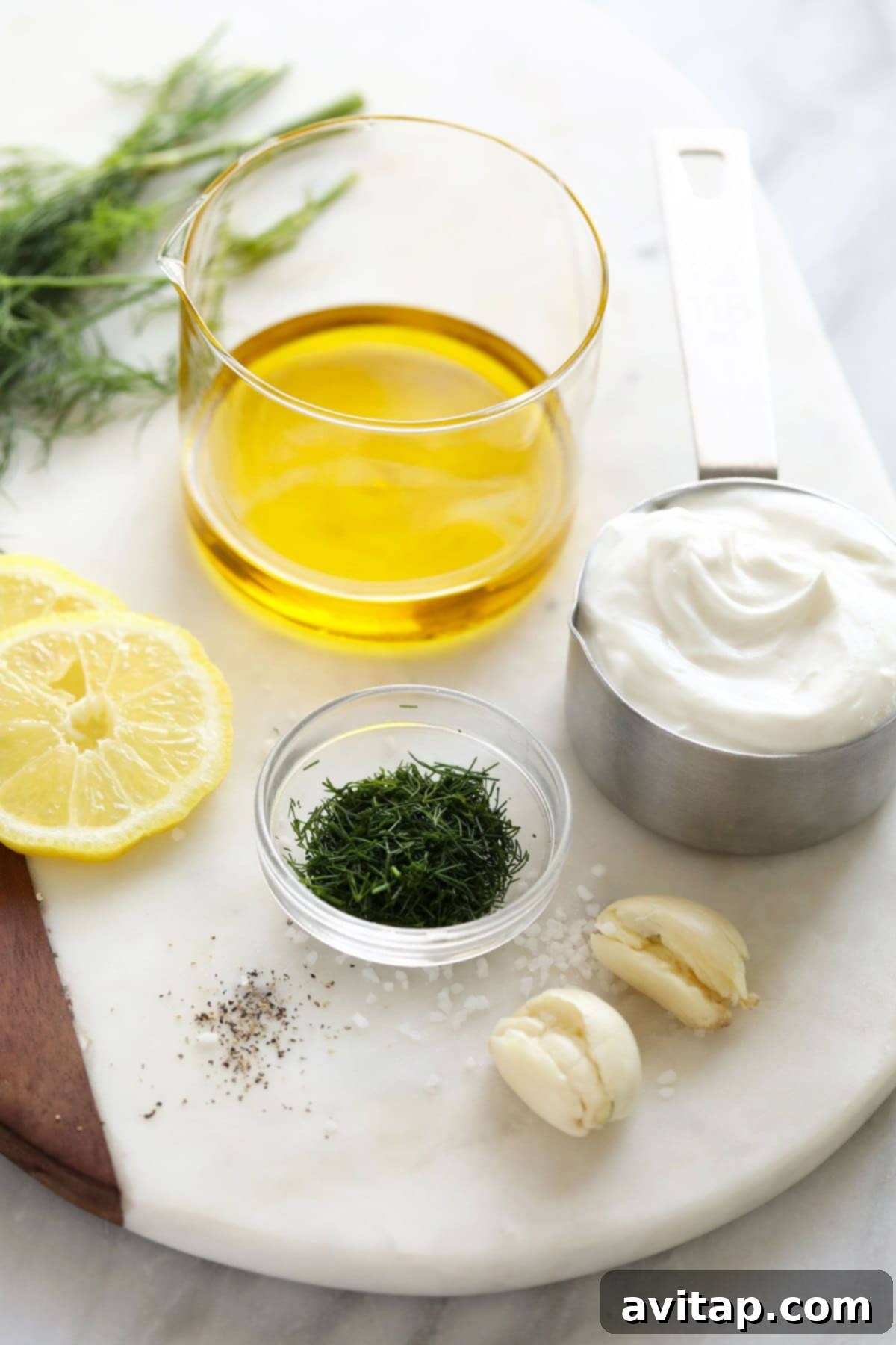 Assortment of fresh ingredients for Greek yogurt salad dressing including dill, lemons, and garlic, arranged on a rustic wooden cutting board.