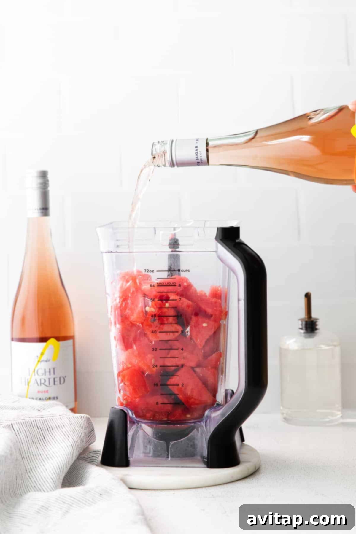 A bottle of rosé wine being poured into a high-speed blender filled with frozen watermelon chunks, ready for blending.
