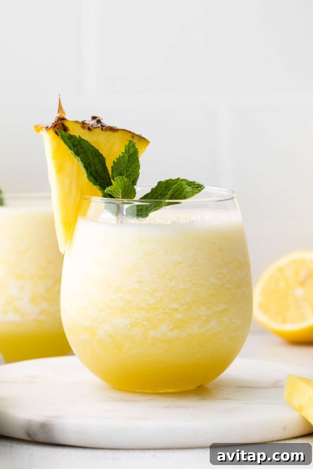 A tall glass of sunny yellow tropical wine slushie, garnished with a fresh pineapple wedge and a sprig of mint.