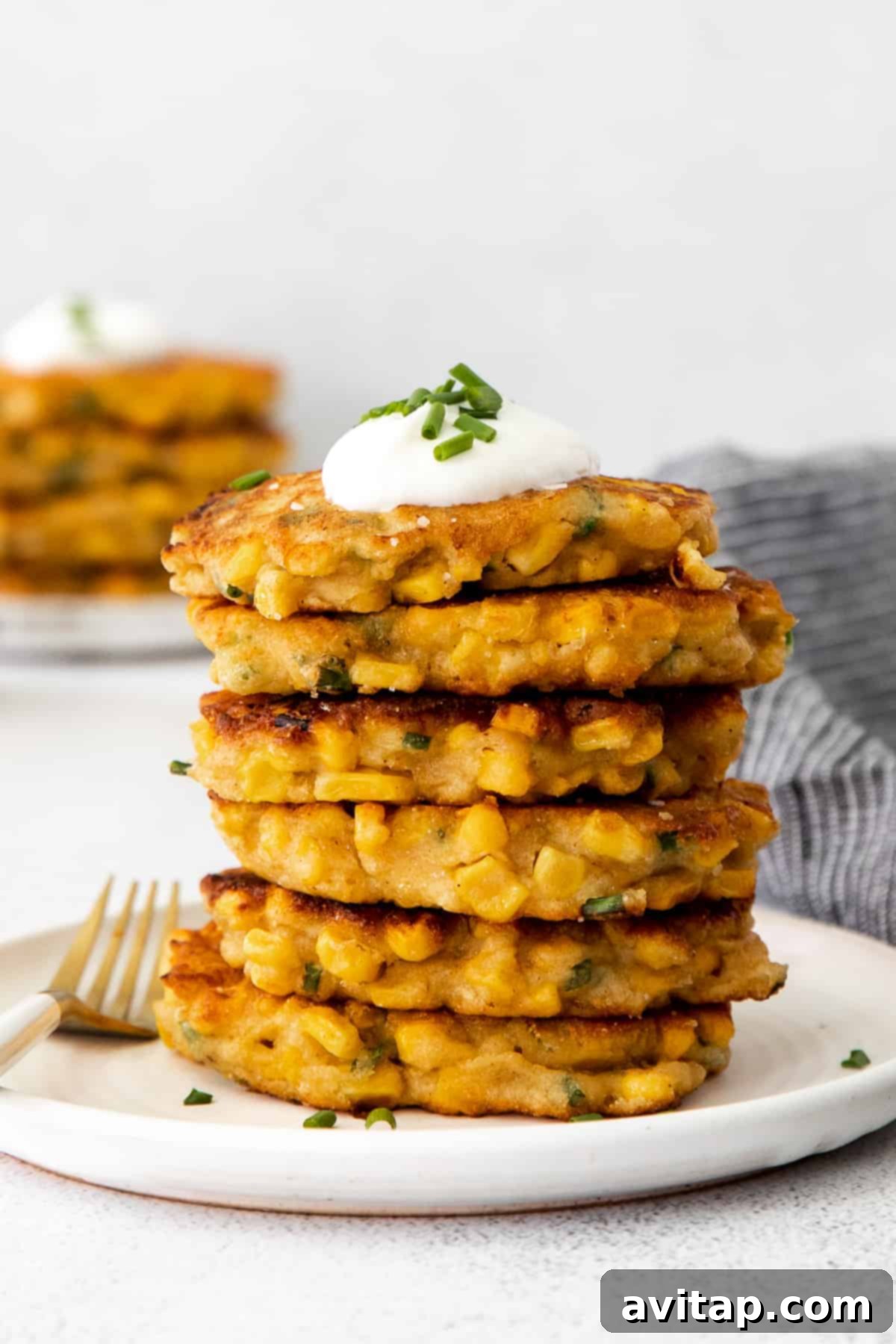 A stack of golden-brown corn fritters topped with a dollop of Greek yogurt and freshly chopped chives, artfully arranged on a plate.