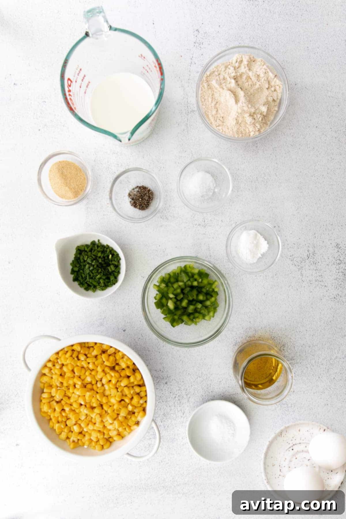 A vibrant collection of fresh ingredients for corn fritters, including golden corn kernels, bright green chives, sliced pickled jalapenos, a bottle of olive oil, and various spices, laid out on a clean surface.