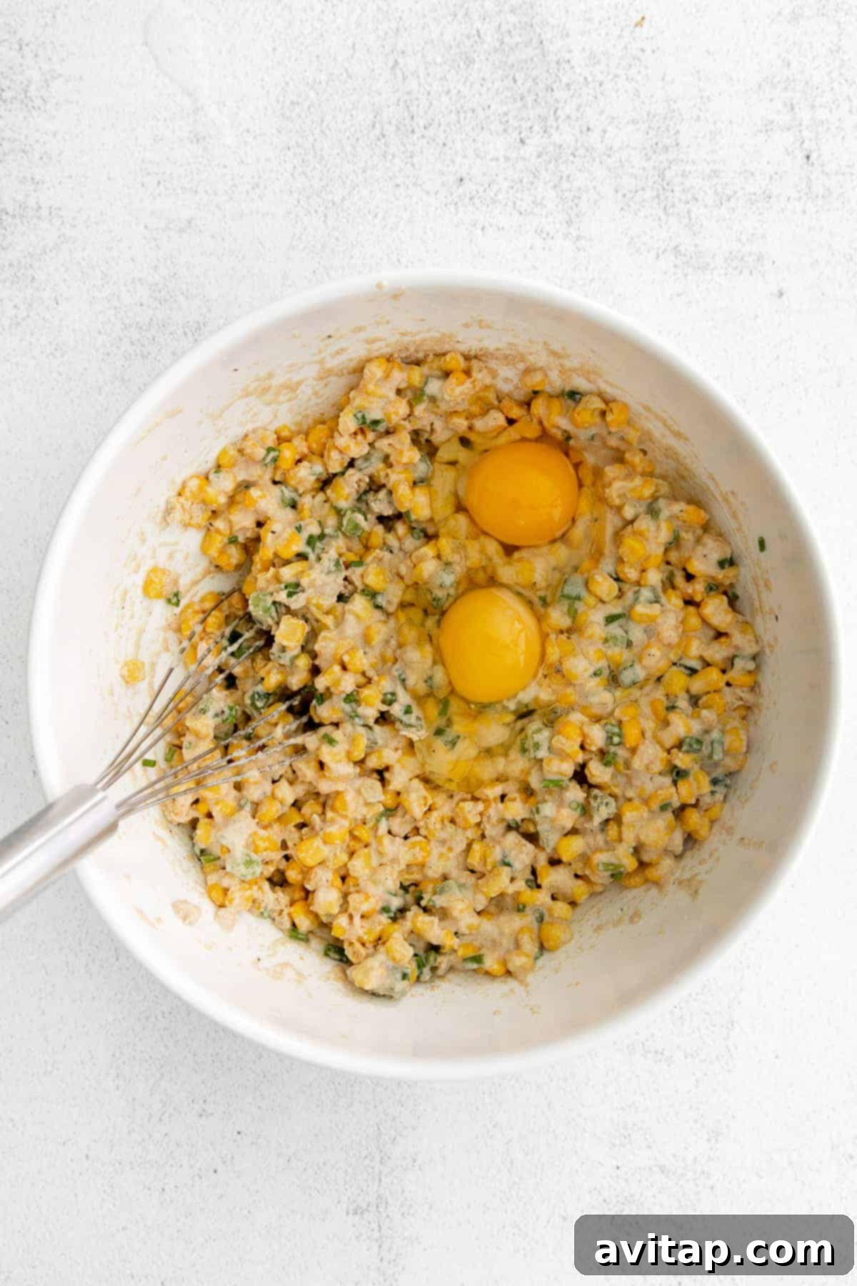 Close-up shot of corn fritter batter in a bowl, with two raw large eggs cracked directly on top, illustrating the mixing process.