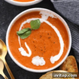 A bowl of creamy, Healthy Tomato Bisque Soup garnished with a dollop of cream and fresh basil, accompanied by slices of bread and golden spoons on the side.