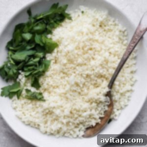 A bowl of perfectly cooked white cauliflower rice, garnished with fresh parsley and a spoon.