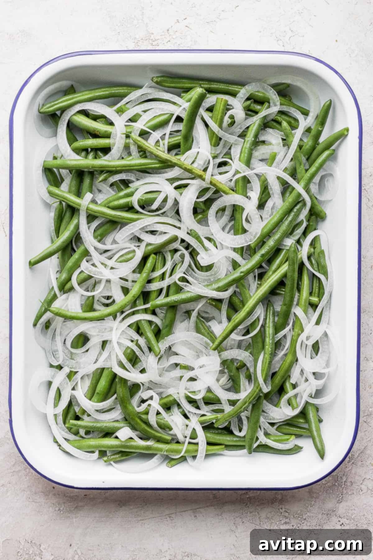 Fresh green beans and sliced white onions carefully arranged in a casserole dish before baking.