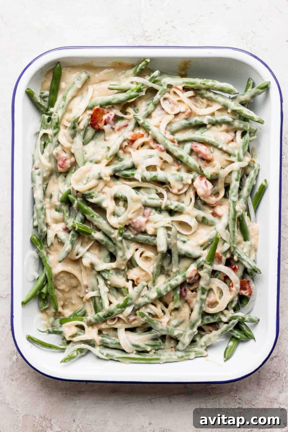A casserole dish filled with green beans, onions, and bacon, coated in a creamy sauce, mixed and ready for the oven.