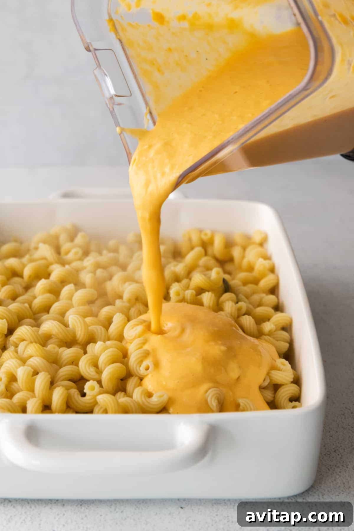 Creamy butternut squash cheese sauce being generously poured over cooked cavatappi noodles in a large bowl.
