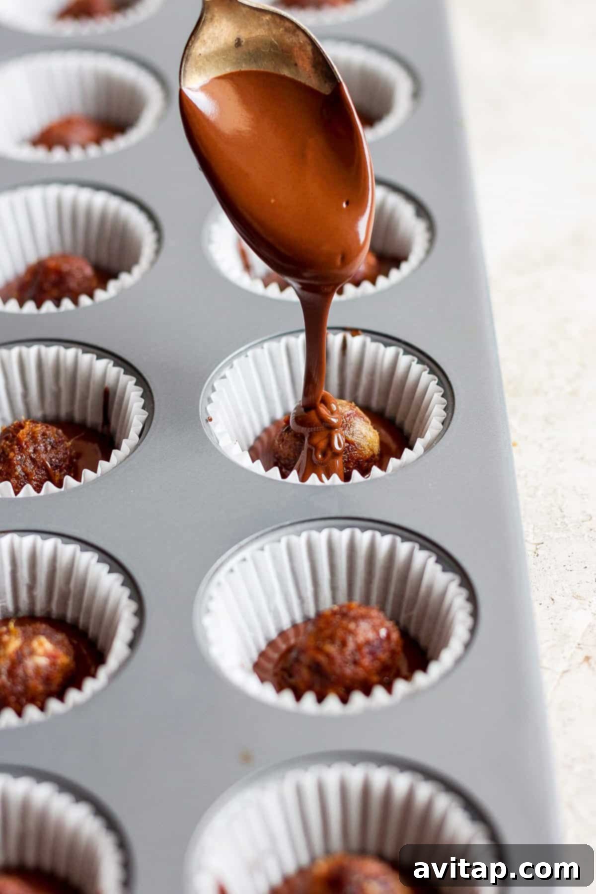 A spoon carefully drizzling melted chocolate over date caramel balls nestled in cupcake liners within a muffin form, demonstrating the coating process.