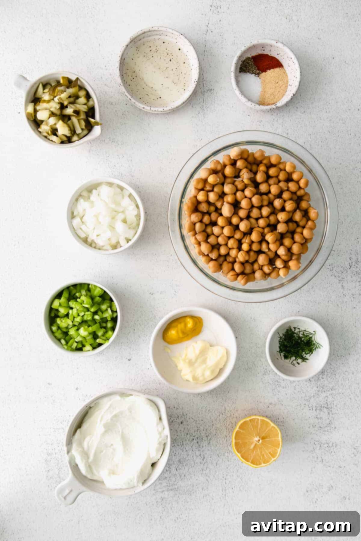 Fresh ingredients laid out neatly on a kitchen counter, showcasing canned chickpeas, finely chopped celery, onion, pickles, and the various components for the creamy dressing.