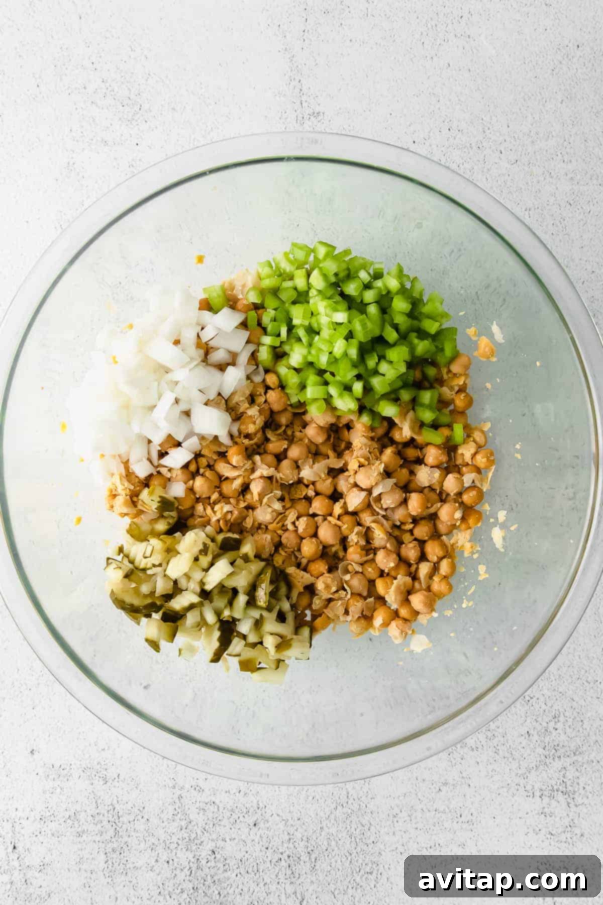 All prepared ingredients for chickpea salad, including mashed chickpeas, chopped vegetables, and the creamy dressing components, ready to be combined in a large mixing bowl.